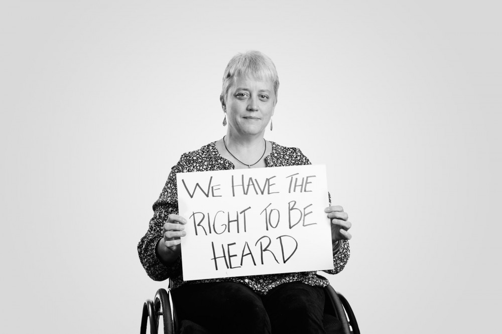 Louise, who uses a wheelchair, holds a handwritten sign: We Have the Right to Be Heard